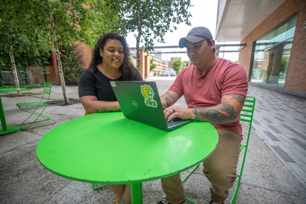 Young female and male students are seated at a green table outside in a campus common space, viewing a laptop screen together.