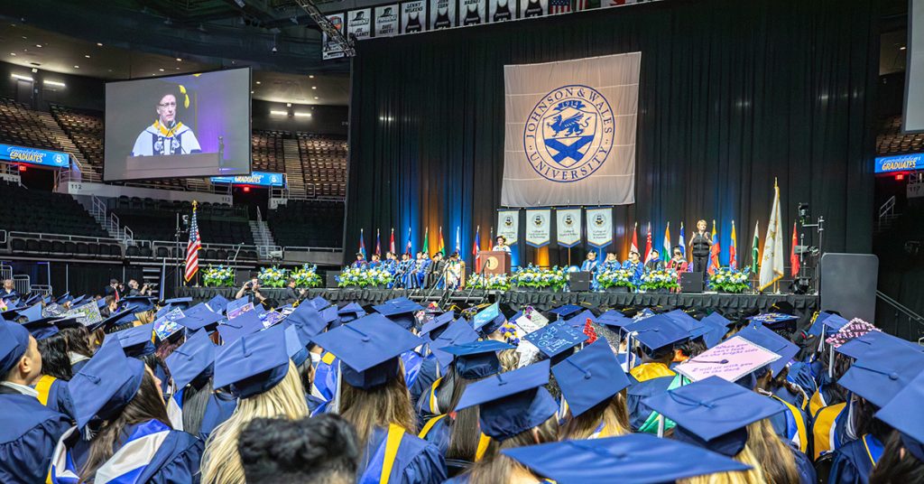 View from behind students in caps and gowns, seated in the Amica Pavilion arena at commencement, as they wait to walk across the stage to receive their diplomas.