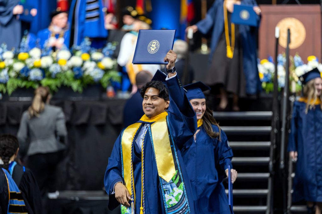 A young male in cap and gown hold his diploma over his head as he exits the stage at commencement,