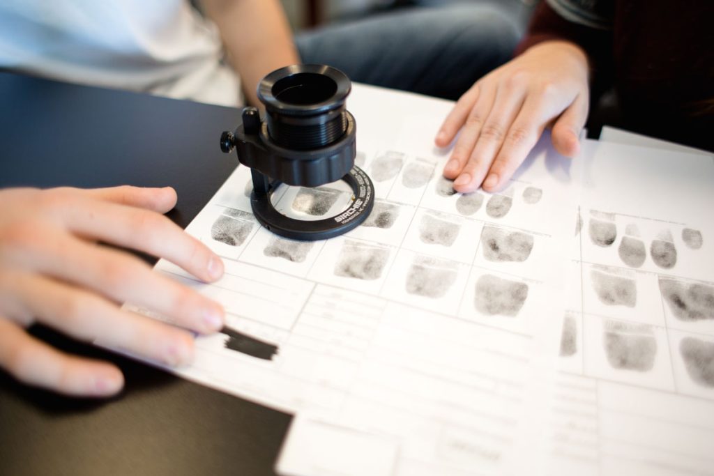 Hands of two different people resting on pages of finger prints on which a fingerprint magnifier (loupe) is positioned for viewing.