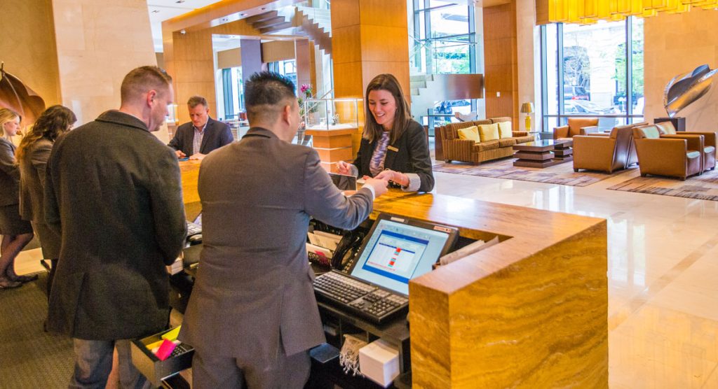 Young woman in a hotel lobby checking in with with a gentleman wearing a suit at the front desk, while a second man looks on.