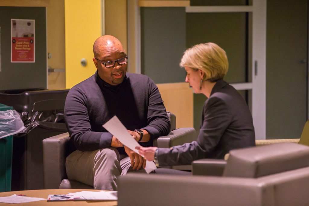 A seated male graduate student and a female staff member, who is holding a document, are talking together.
