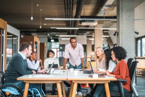 A diverse team of colleagues gathered around a table in a modern office, discussing brand strategy together with laptops and notebooks open.