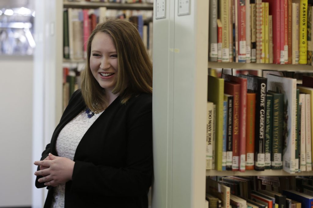 A young woman with a big smile , stands in front of shelves lined with books in the Johnson & Wales University library.