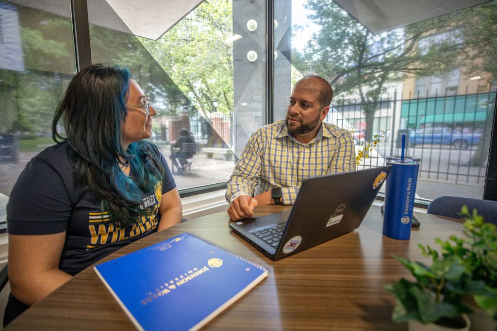 A male staff member seated with a laptop talks with a student seated next to him in an office with windows behind them facing out to the street.