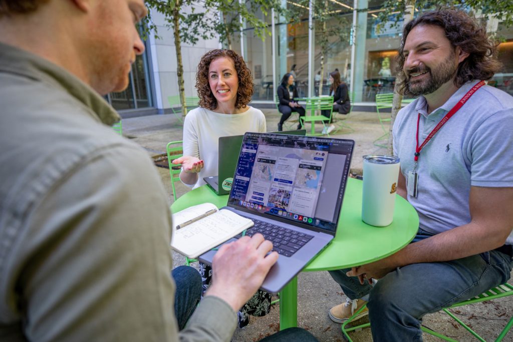 Two male students, one looking at his laptop screen, and a female student are seated at a cafe table in a common area outside the Bowen Center on the Providence Downcity Campus.