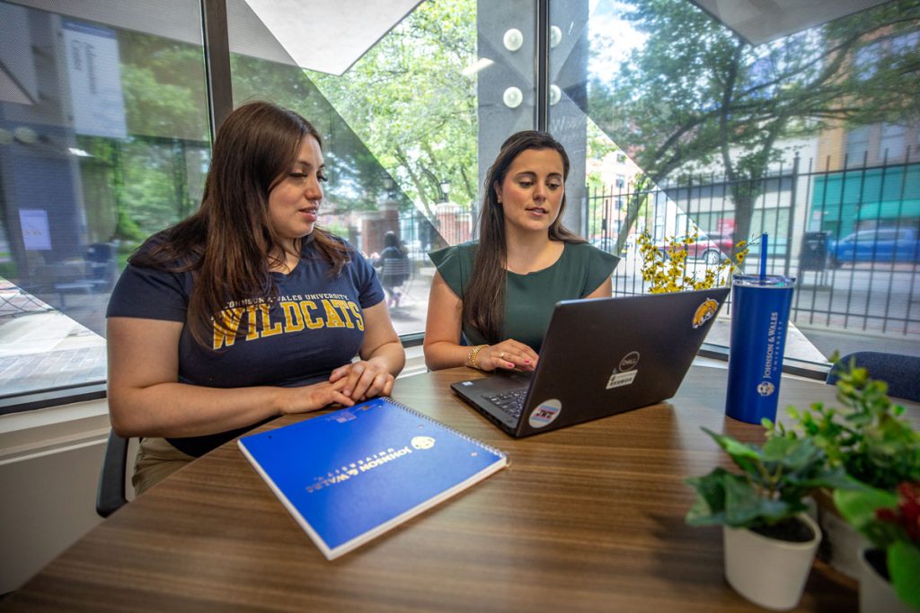 A young woman wearing a Wildcats T-short is seated next to a woman with a laptop in front of windows in a JWU office that look out on the campus and city street.