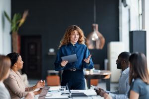 Woman holding a tablet leads a team meeting with four colleagues seated at a conference table in a modern office