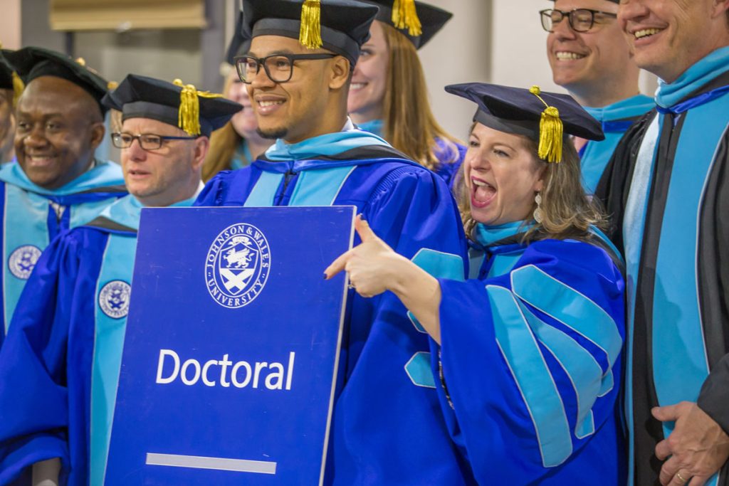 A group of male and female students wearing royal blue doctoral gowns with light blue velvet panels are displaying a sign with the JWU seal and the word "Doctoral," to which one woman is pointing.