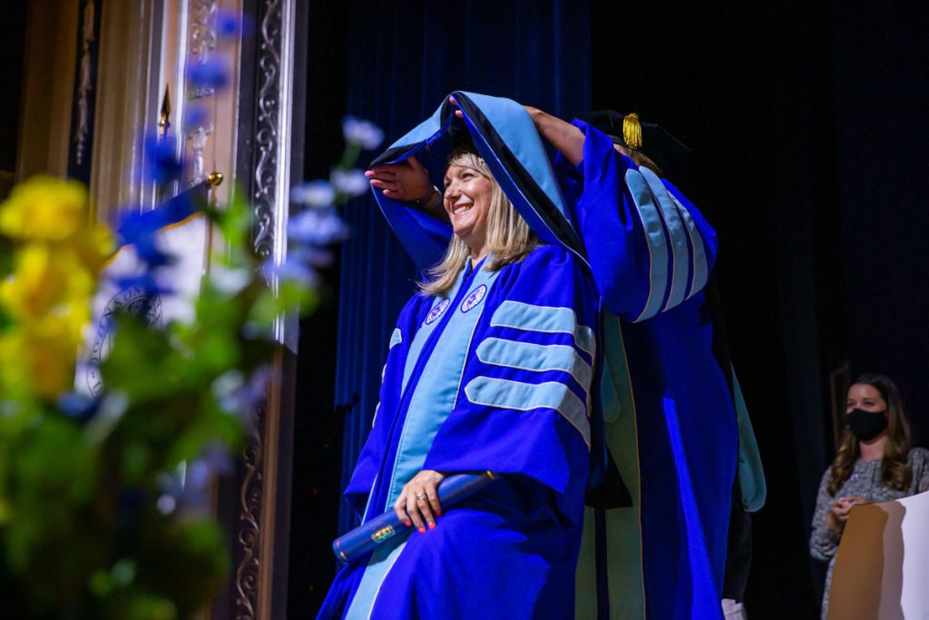 Smiling woman with blonde hair wearing a royal blue doctoral gown with light blue velvet panels as she is ceremonially draped with an academic hood by a faculty member during a JWU commencement