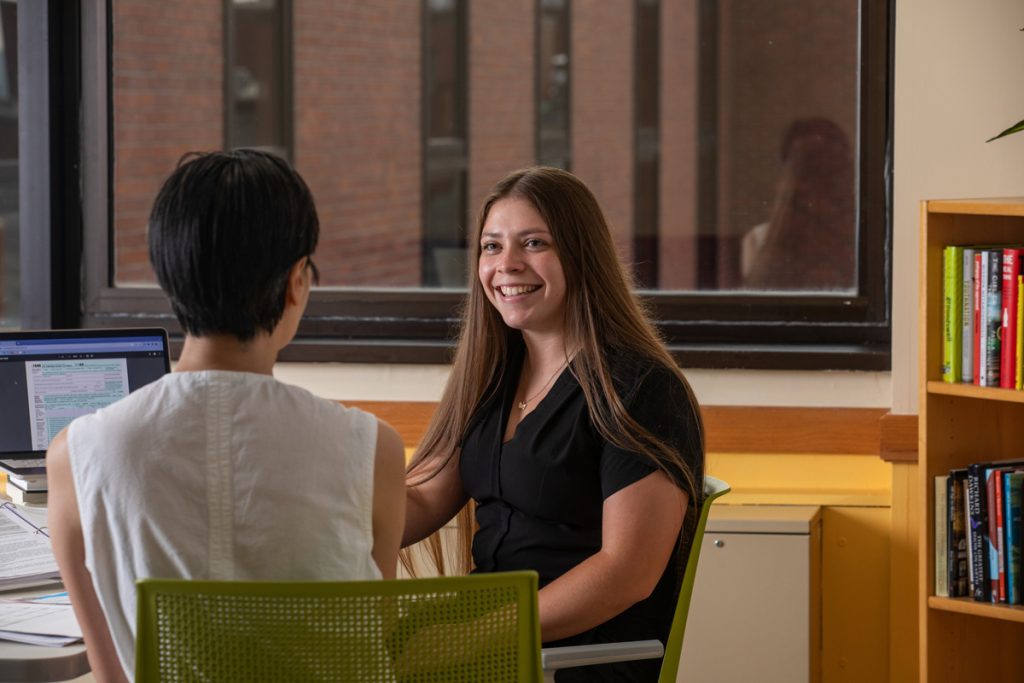 A young woman is smiling as she faces a female staff member she is meeting with for assistance in an office in a JWU campus building.