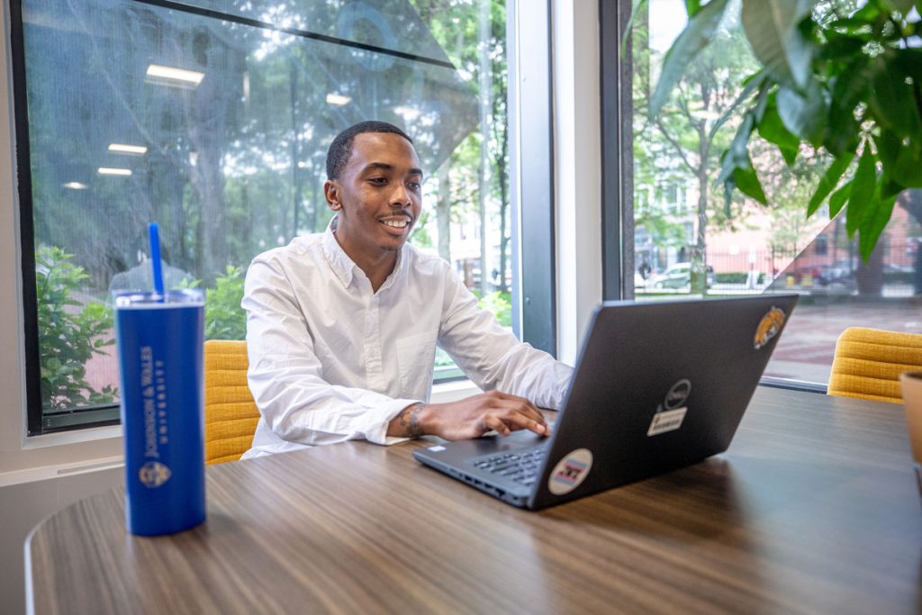 Young man seated at a table, working on a laptop, in an area with windows behind him that look out on a campus courtyard at the JWU Downcity campus in Providence, RI.