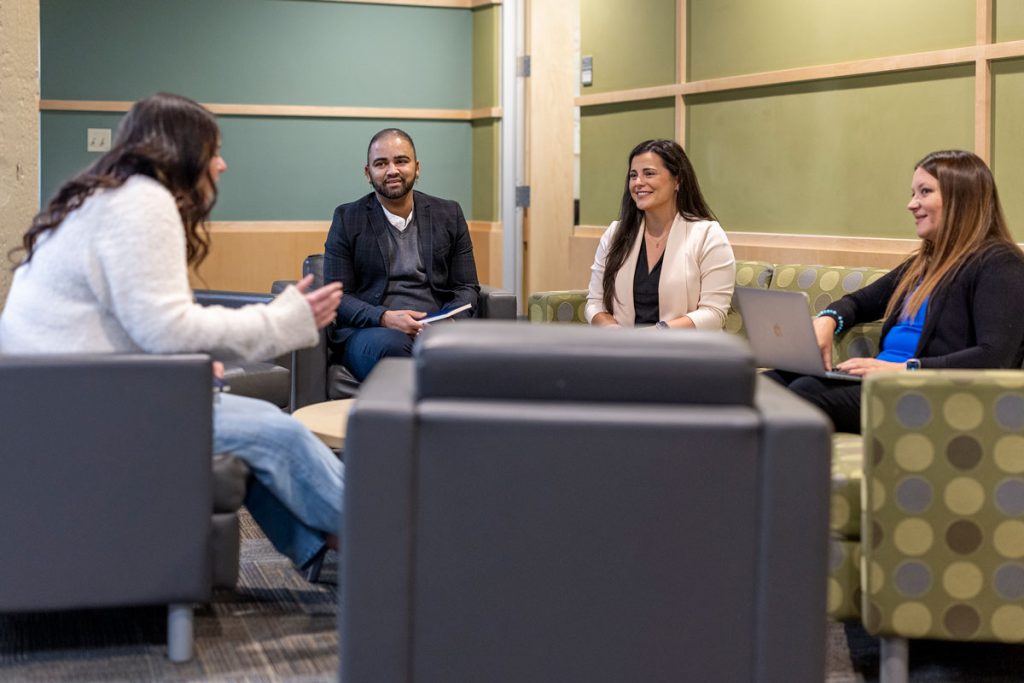 Three young women and one young man, all dressed casually, are seated in a lounge area, talking with one another.