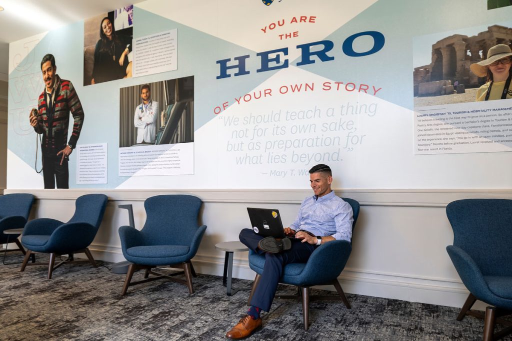 Young man in business casual clothing, seated in a blue chair and working on his laptop, while behind him is a wall with a mural on which is lettered: "You are the hero of your own story."