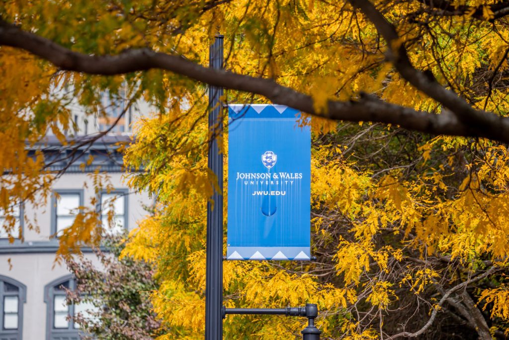 A blue banner for Johnson & Wales University mounted on a pole, with vibrant yellow autumn foliage and a campus building in the background.