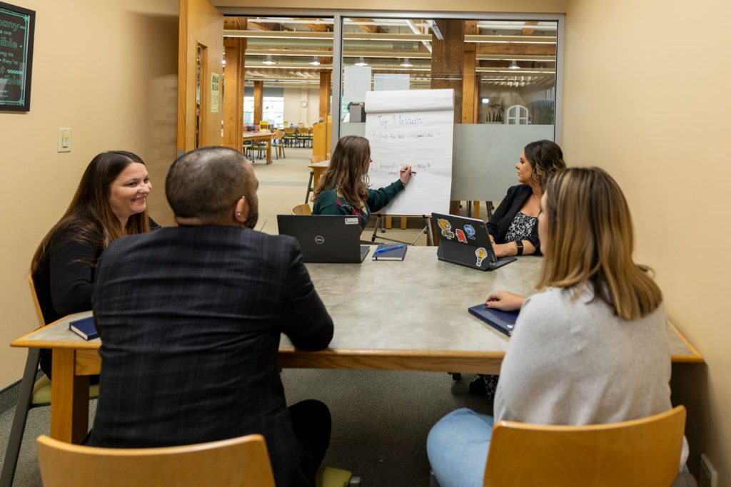 Three female and one male seated at a table in the Friedman Center library are listening to a young woman who is writing on a large pad as she speaks.