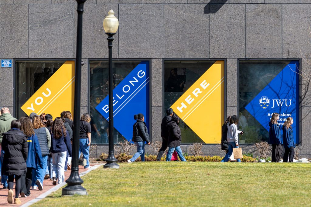 Students walking towards the JWU Providence Admissions Center at Gaebe Commons on which gold and blue banners display, "You Belong Here" and the JWU logo.