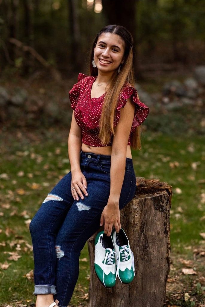 Photo of Peyton Nardella, smiling while seated on a tree stump in a wooded outdoor area, wearing a red crop top, blue jeans, and holding a pair of white-and-green sneakers.