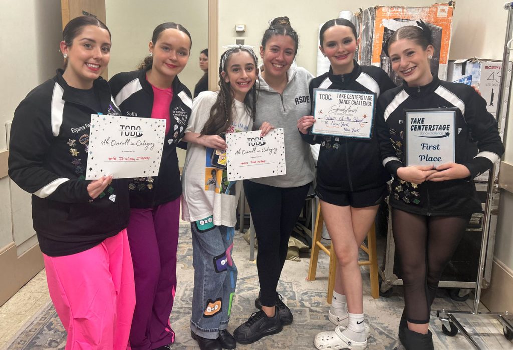 Six young women who are students in the dance class Peyton Nardella teaches are smiling while they hold certificates showing their achievements in dance competitions.