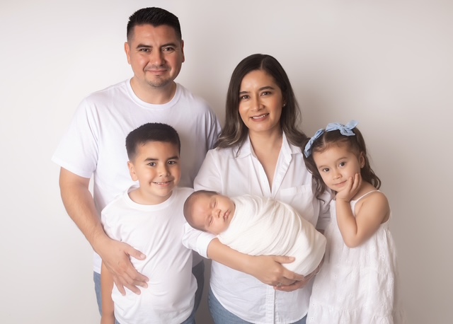 Carlos Coronado standing with his wife, who is holding their baby son, his daughter, and older son, all wearing white tops, against a light background.