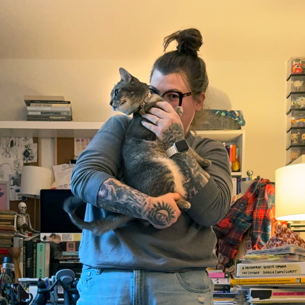 Mikala Schimke in her home office with her face half-hidden by the cat, Houdini, that she is holding.