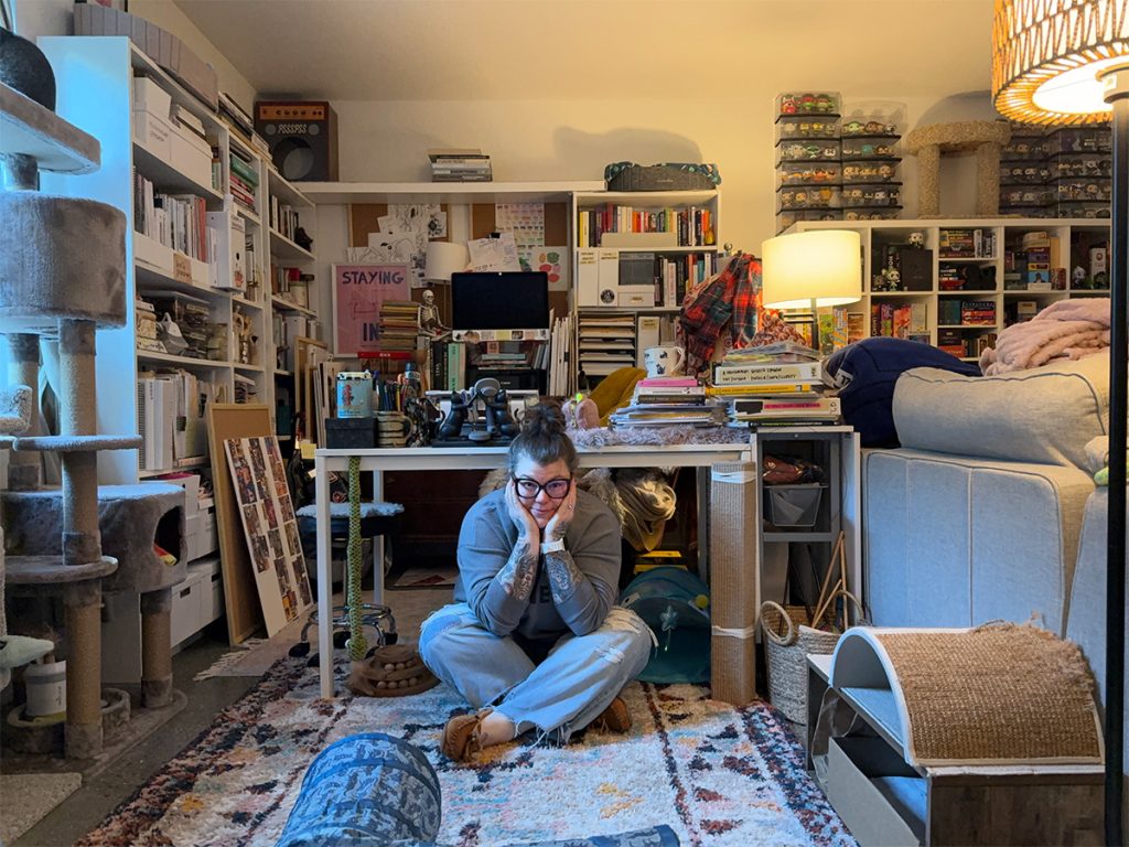 Mikala Schimke sits on the floor, cross-legged with chin resting in hands on either side of her face, surrounded by shelves holding craft supplies in her home office.