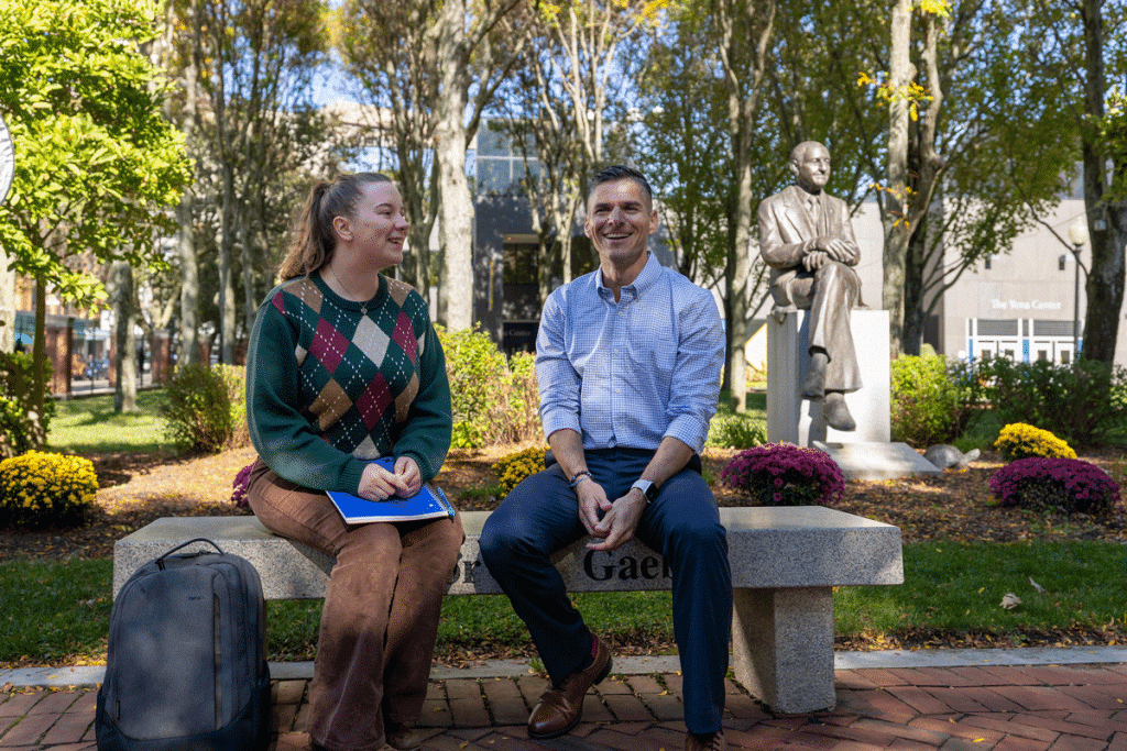 Female and male dressed casually, talking together while seated on a bench in Gaebe Commons, on the Providence Downcity JWU campus.